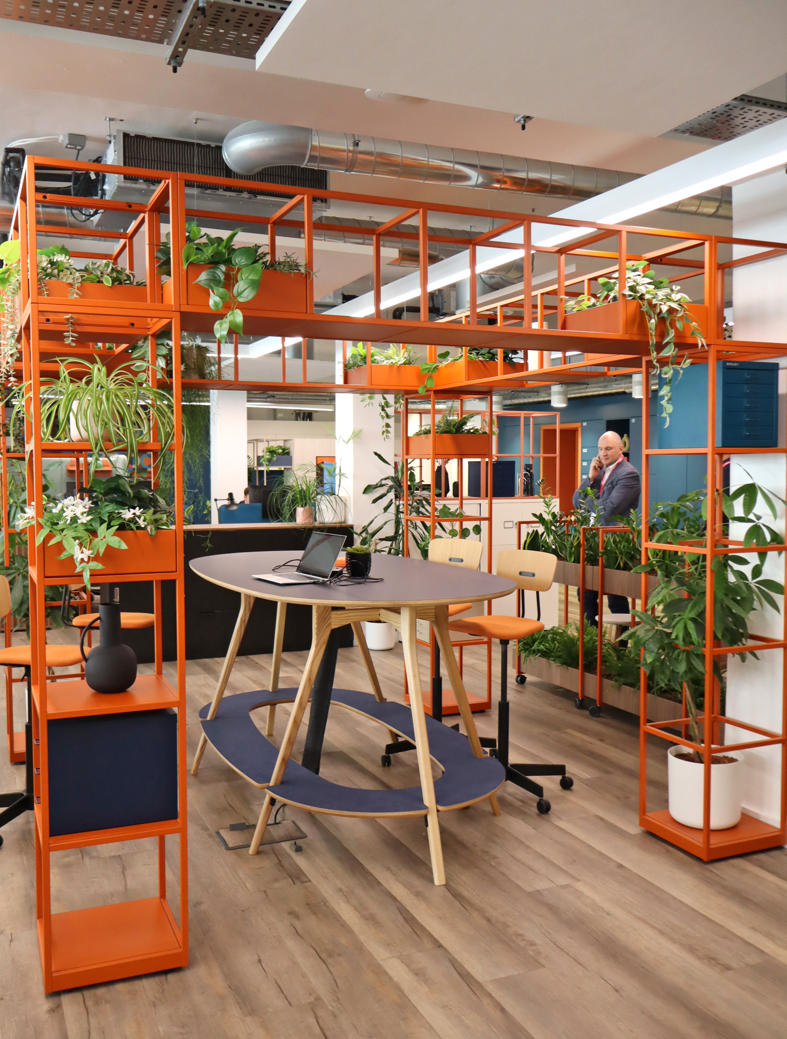 a poseur height desk, surrounded by steel frame storage. lots of colour on the furniture, oranges, blues, and lots of plants sitting on and hanging from storage.